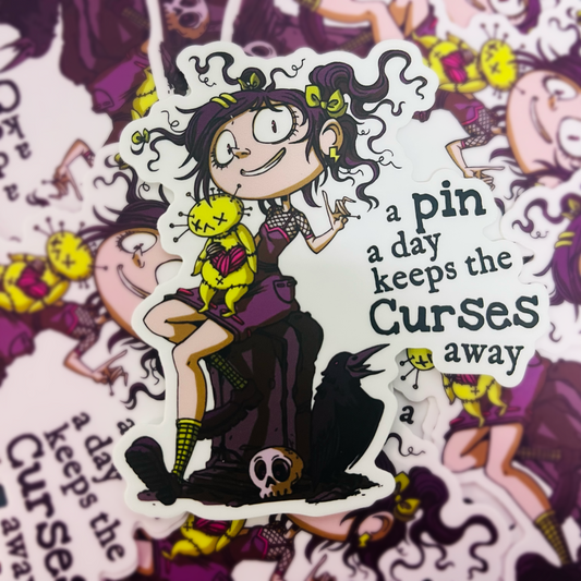A vinyl sticker of a cute girl holding a voodoo doll and sitting on a tombstone and showing a pin. A sentiment saying "a pin a day keeps the curses away" is also on it.